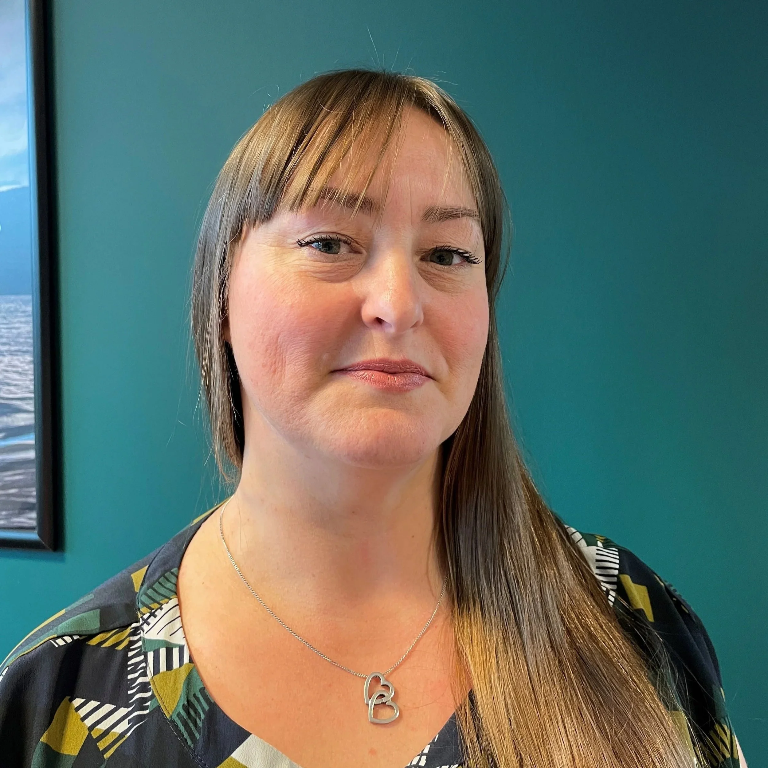 Rebecca is smiling, she has long brown hair with a touch of auburn in it and is wearing  a double heart necklace and autumn colours blouse.