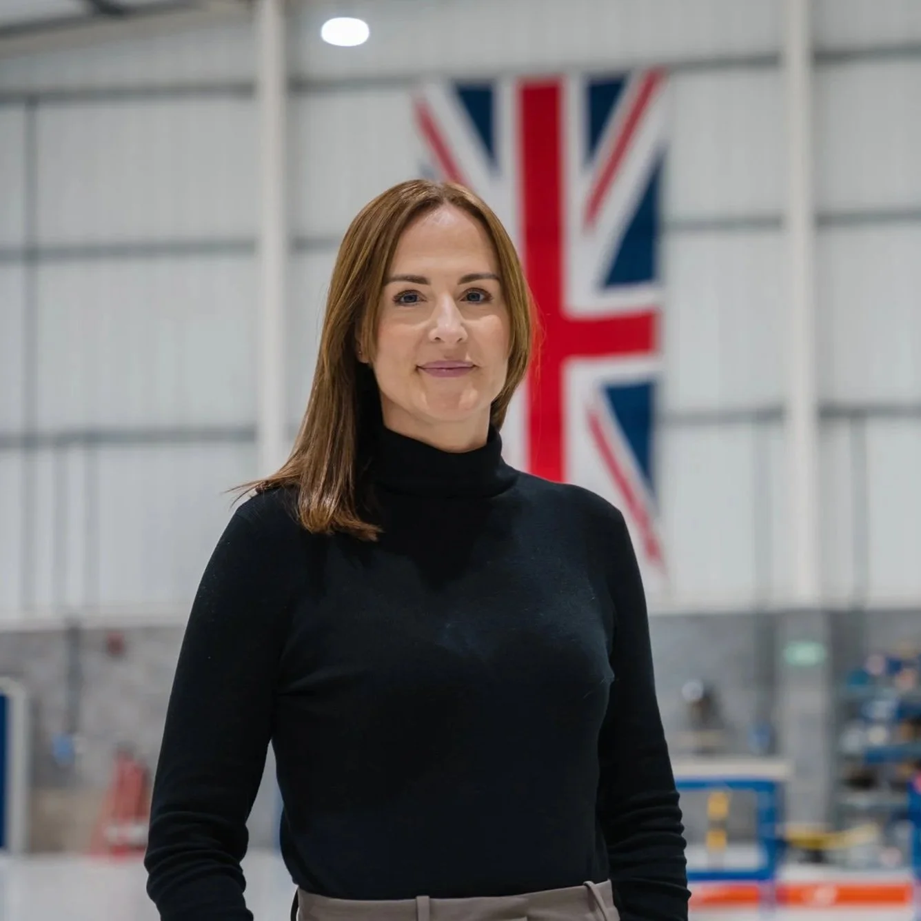 A woman with shoulder-length brown hair, wearing a black turtleneck, standing in front of a large Union Jack flag inside a building.