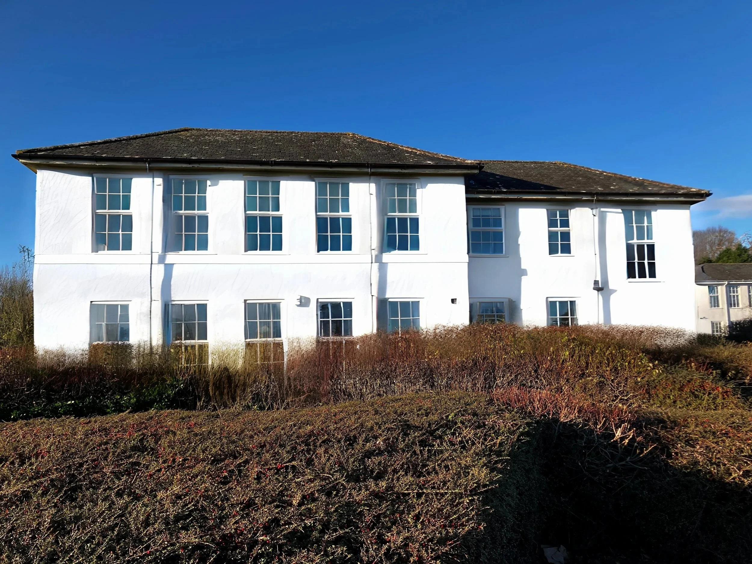 A white multi-story office building with large windows, set against a clear blue sky and surrounded by a garden with bushes and shrubs.