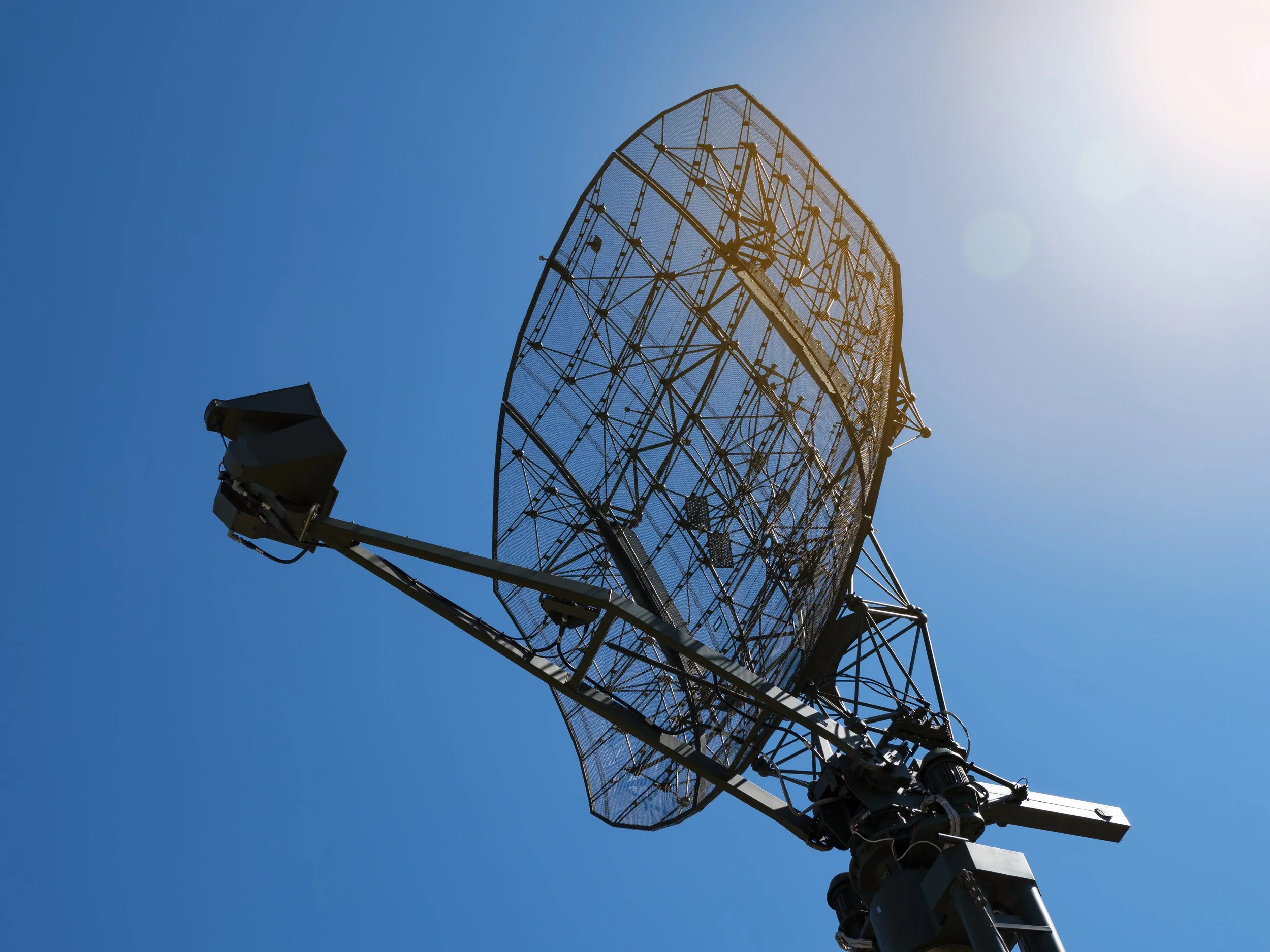 A large satellite dish antenna pointing upward against a clear blue sky with sunlight and lens flare.