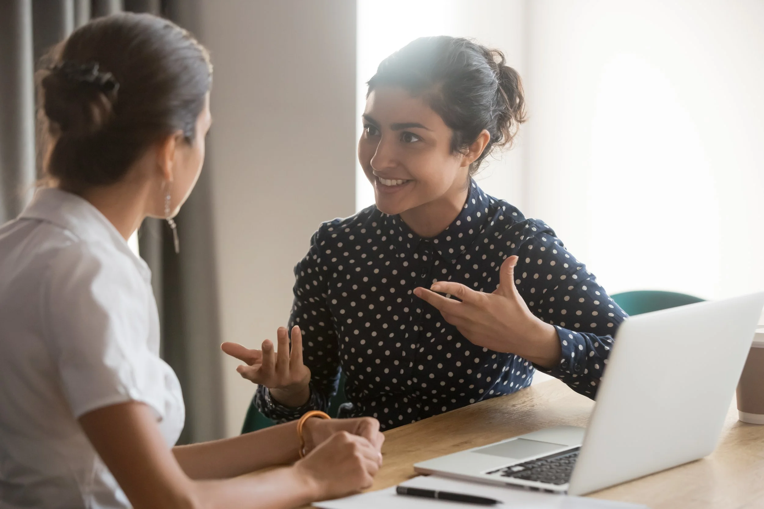 Two women having a discussion at a table, with laptops and a coffee cup, in a bright room.