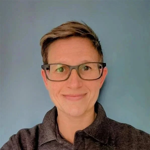 A person with short, styled hair wearing glasses and a dark collared shirt, smiling in front of a light blue background.