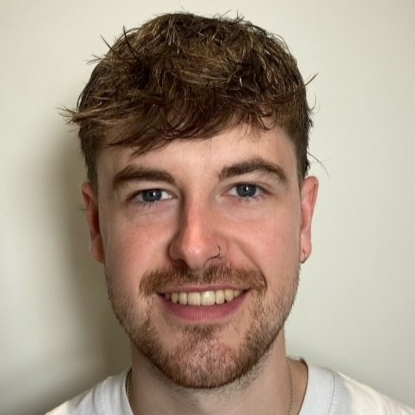Close-up of a young man with brown hair, blue eyes, and a beard, smiling and wearing a white shirt.
