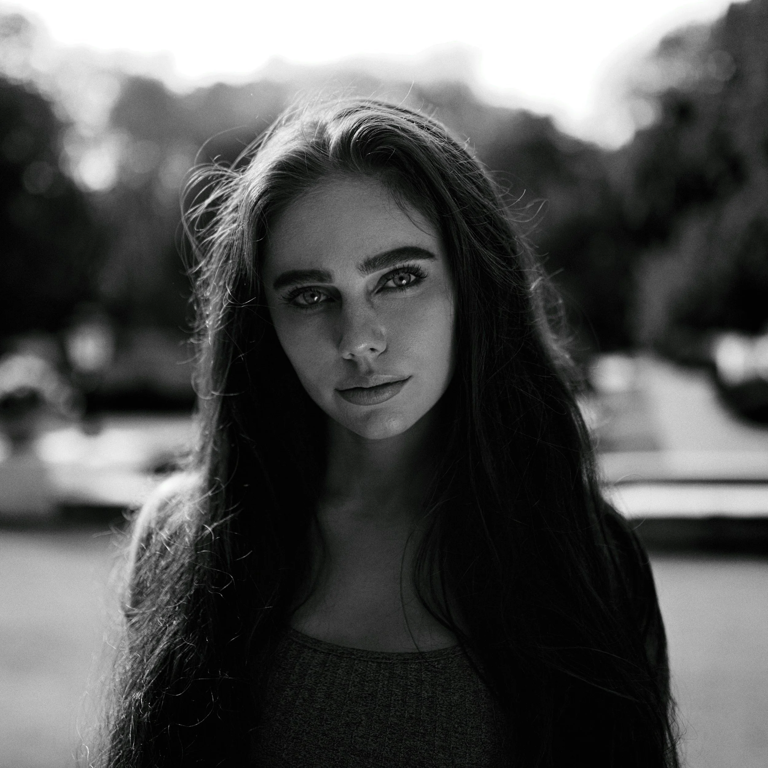 Black and white portrait of a young woman with long dark hair and light eyes, outdoors with blurred trees and cars in the background.