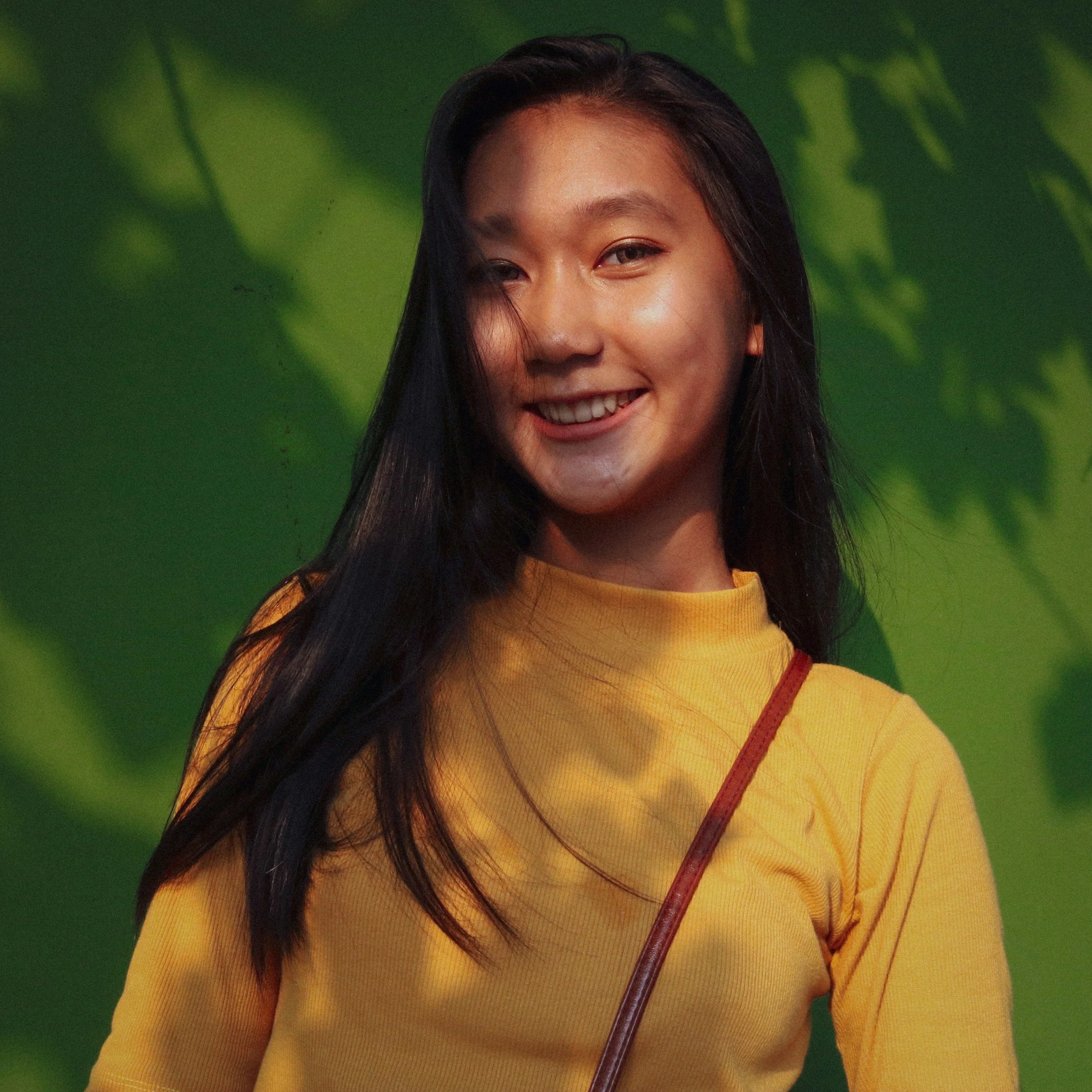 A young woman with long black hair wearing a yellow top and carrying a brown shoulder bag, smiling outdoors with green leaves in the background.