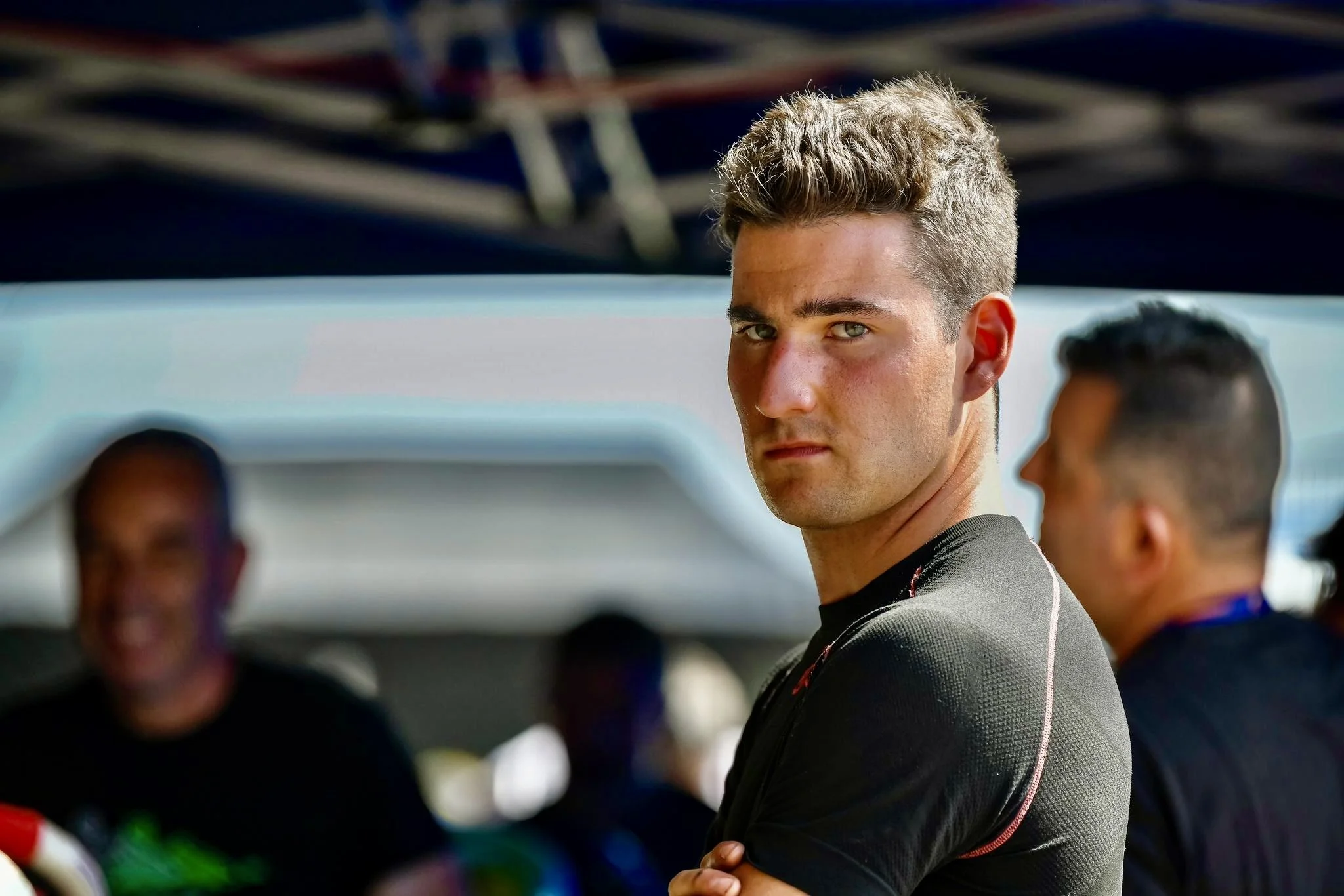 Joven piloto de carreras con miranda seria, de pie en un ambiente de paddock con otros oficiales o mecánicos en el fondo.