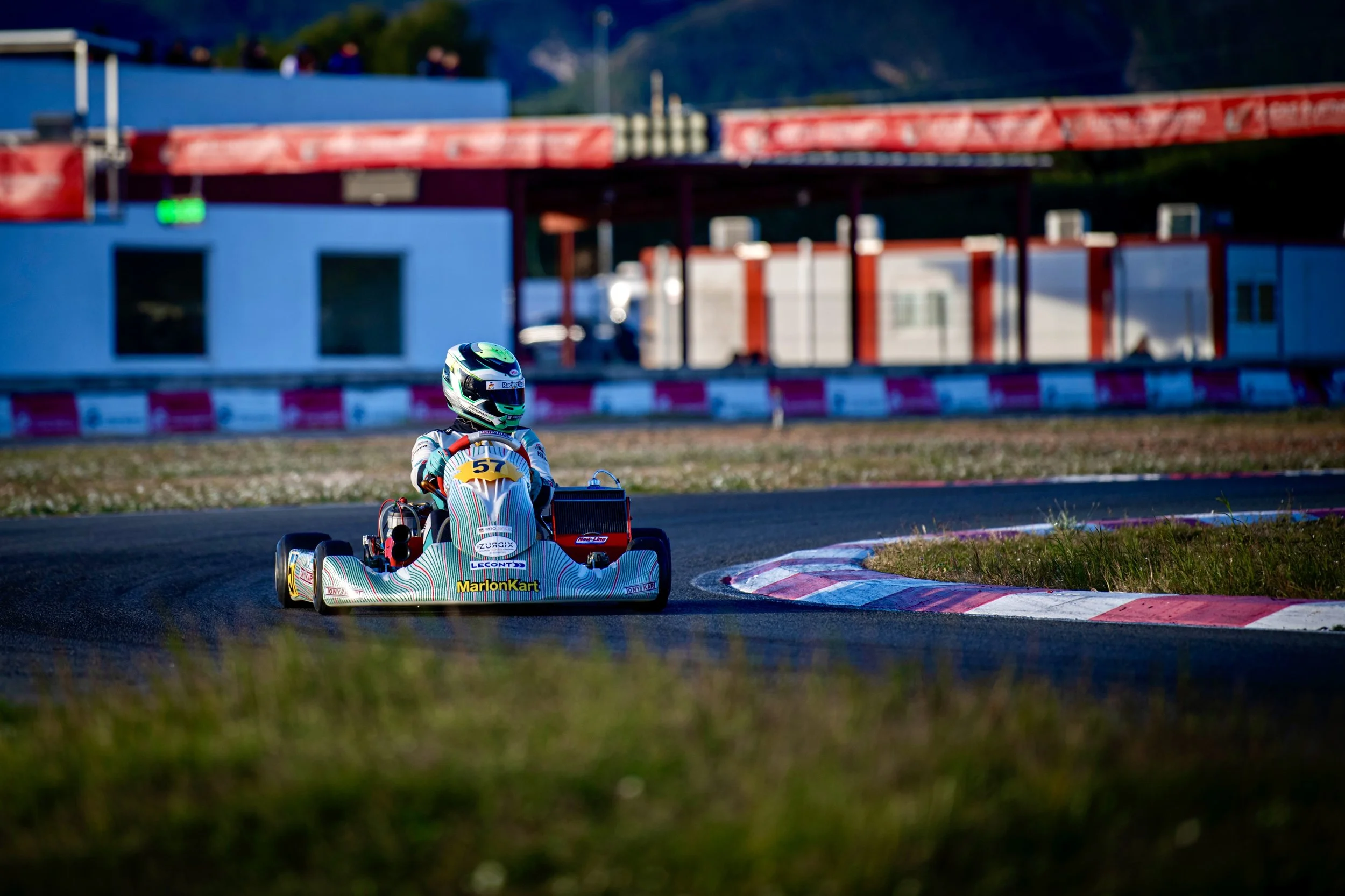 Un niño conduciendo un go-kart en una pista de carreras, con un casco y traje de piloto, en un día soleado.