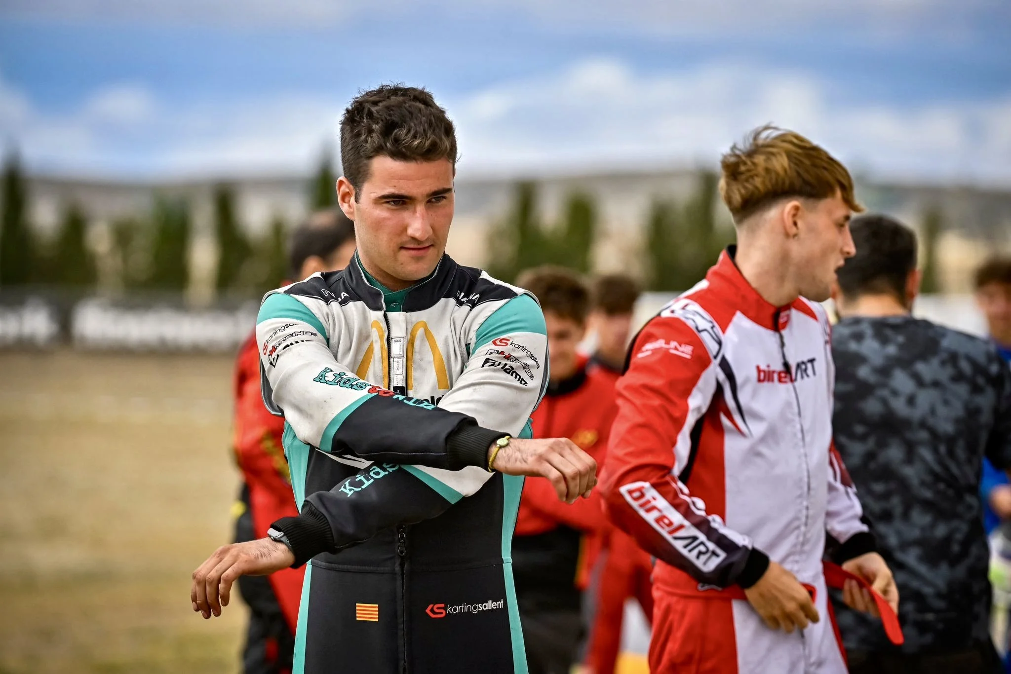 Jóvenes pilotos de carreras en pista, uno con traje azul y otro con traje blanco y rojo, en un entorno al aire libre con cielo y árboles de fondo.