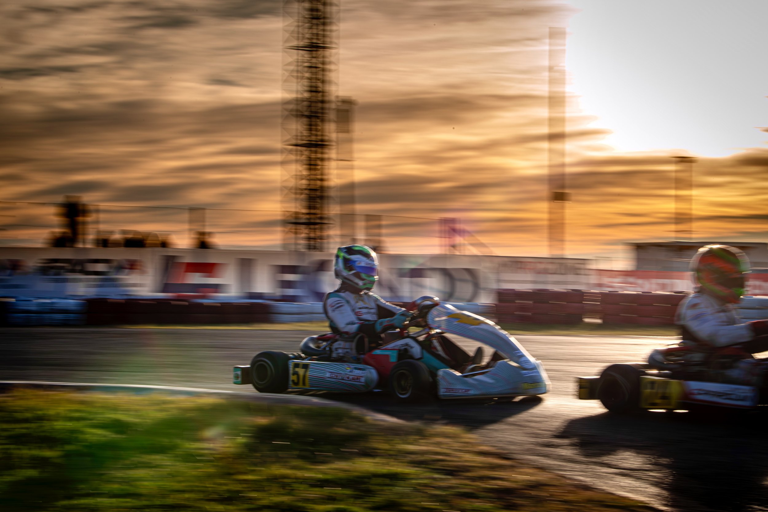 Dos pilotos de kart compiten en una pista durante el atardecer, con una estructura metálica y un cielo nublado en el fondo.