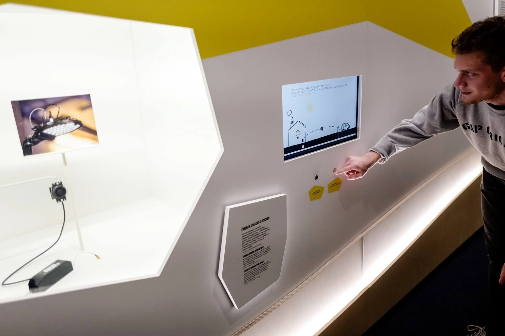 Jeune homme interagissant avec une installation interactive sur un mur blanc, avec un écran numérique et un éclairage LED, montrant une expérience éducative ou scientifique.