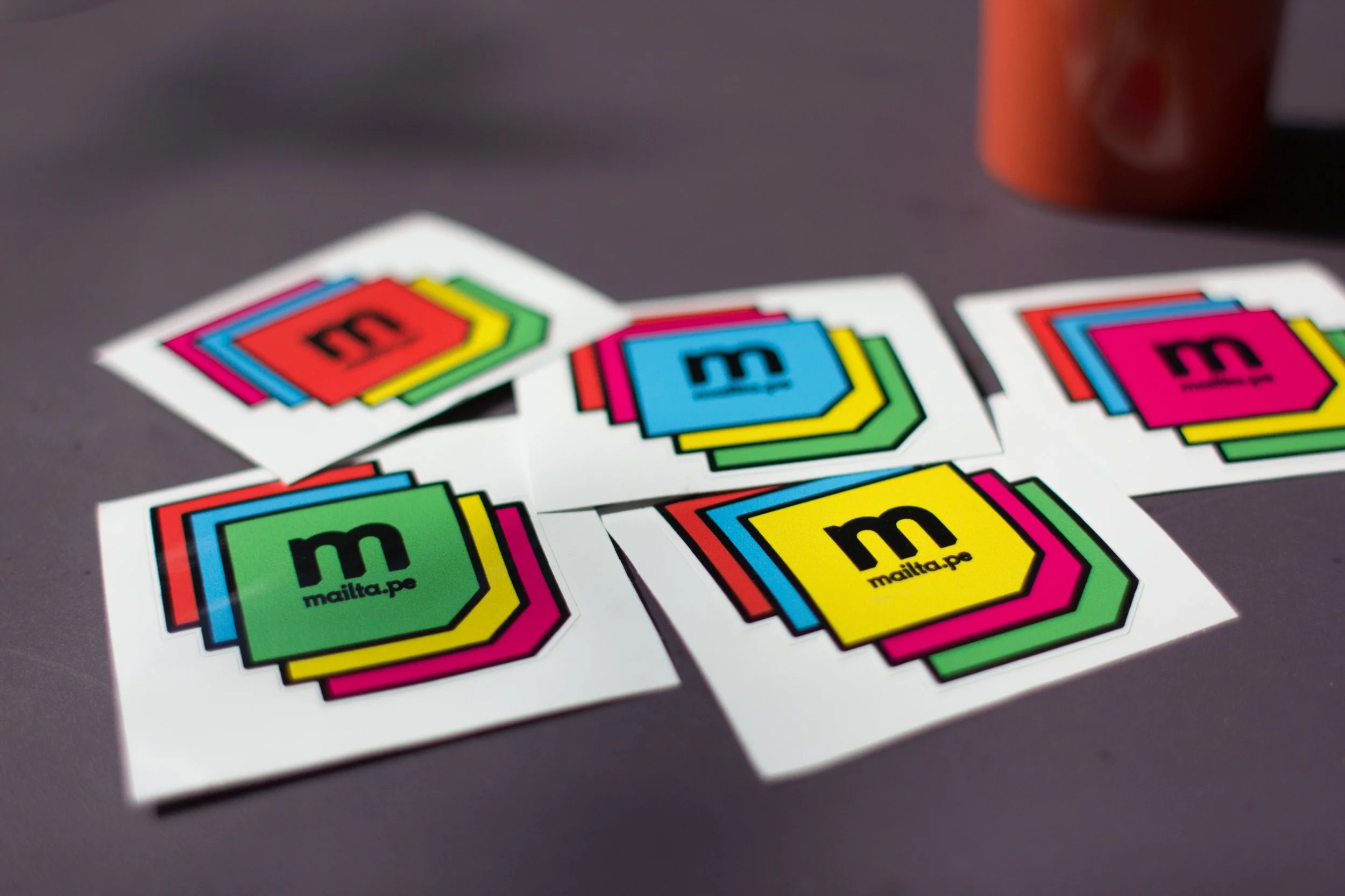 Multiple colorful stickers with the letter 'm' and the text 'mailta.pe' on a gray surface.