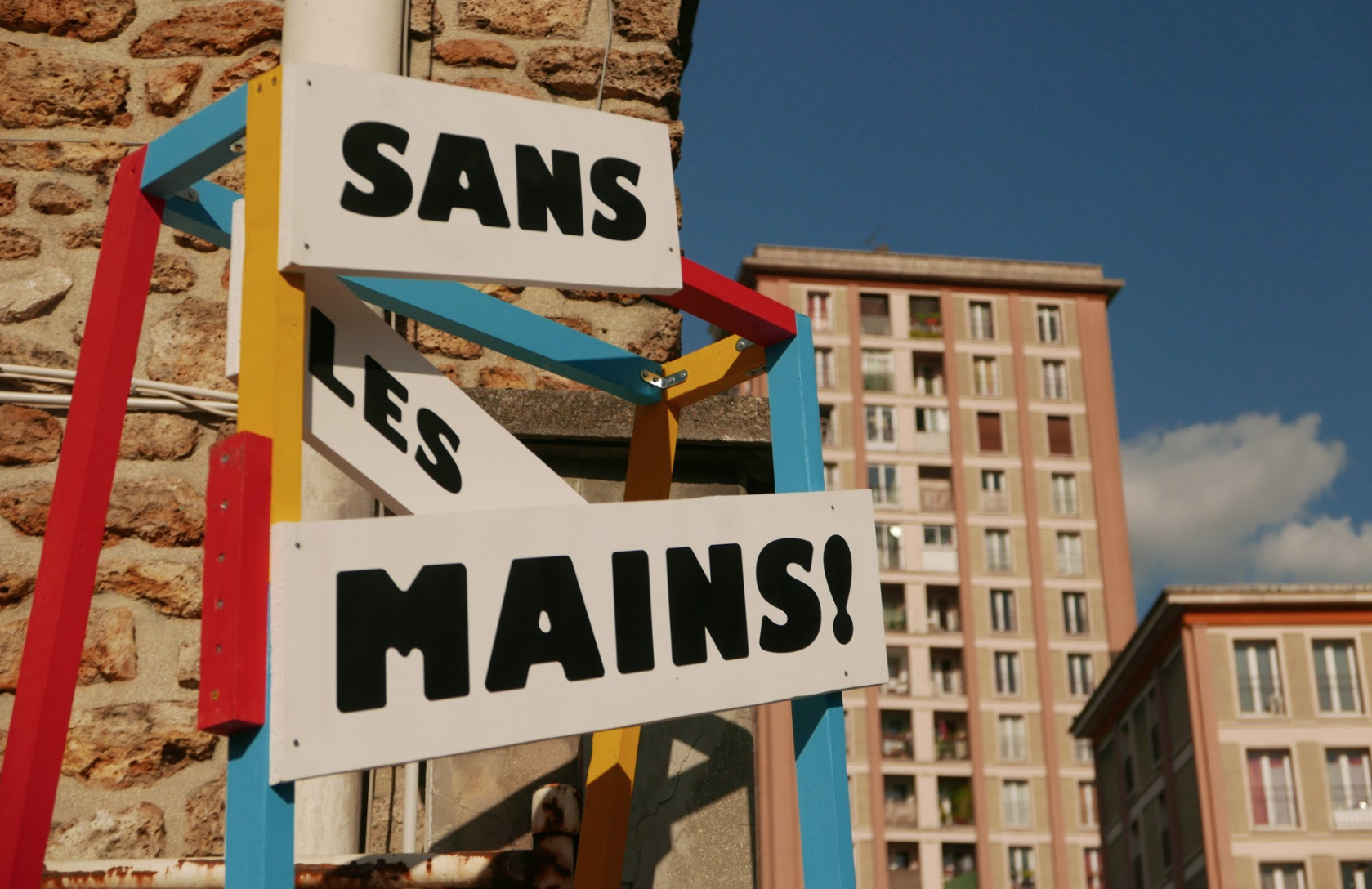 Un panneau coloré avec l'inscription «SANS LES MAINS!» fixé à un mur de briques, avec un immeuble résidentiel en arrière-plan et un ciel bleu. ©GuillaumeBlot