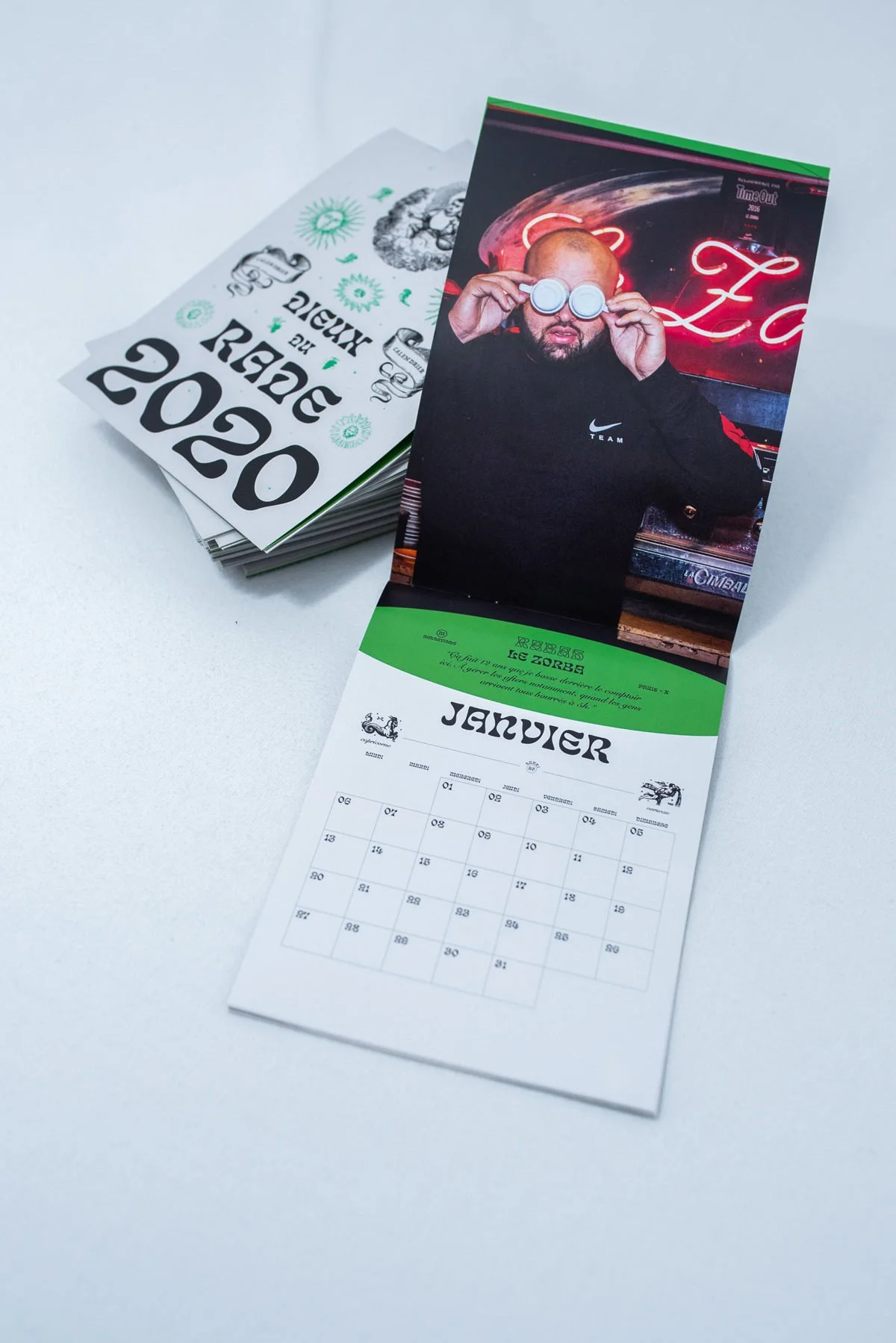 Un calendrier mural ouvert sur le mois de janvier, avec une photo d'une personne portant des lunettes de soleil en arrière-plan. À côté, une pile de calendriers 2020.