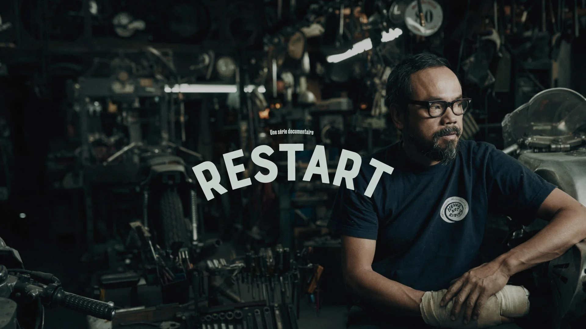Un homme avec des lunettes et une barbe, assis dans un atelier ou un garage avec divers outils et pièces de moto autour, regardant vers la droite. Titre du documentaire 'Restart' en surimpression au centre.