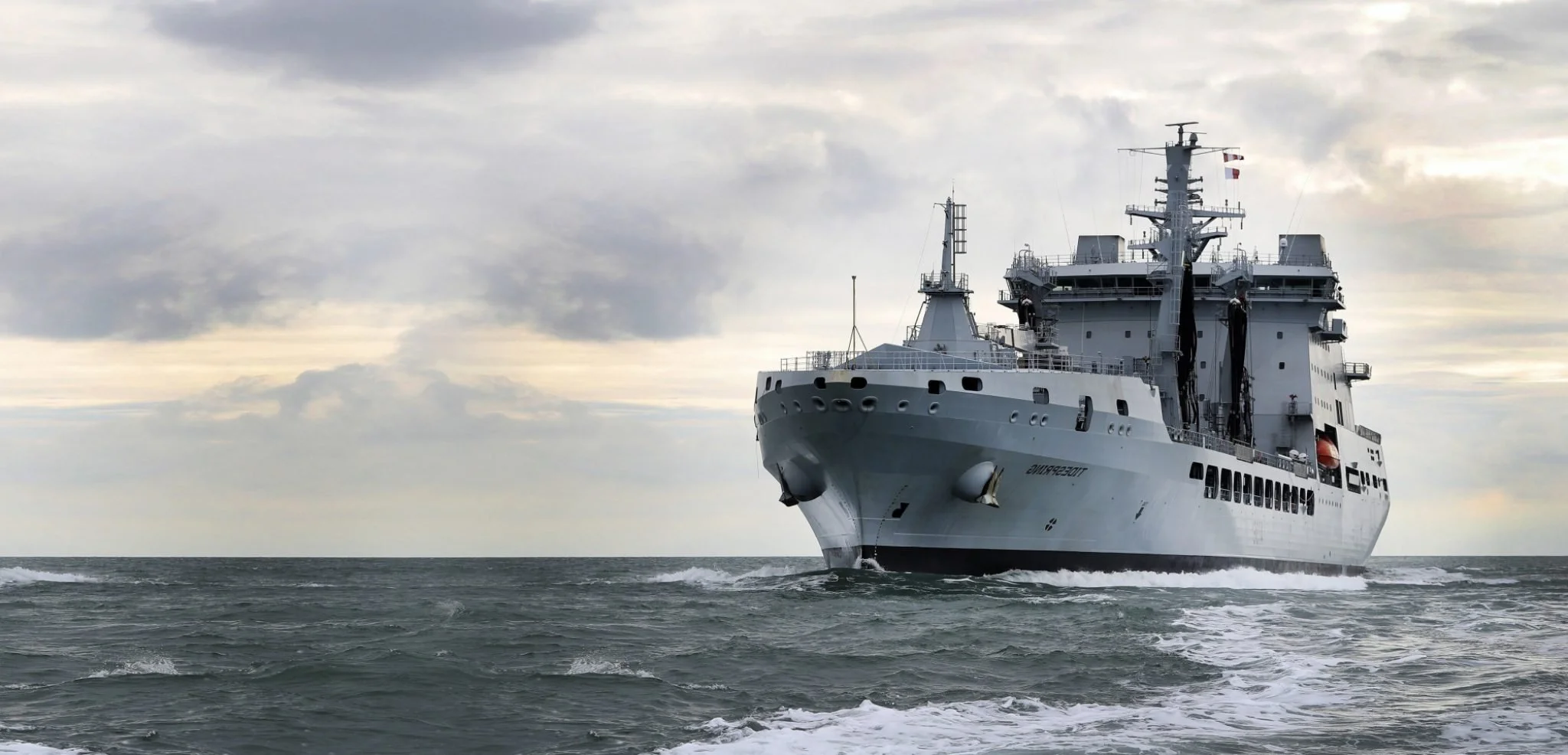 A large gray military ship sailing in the open ocean under cloudy sky.