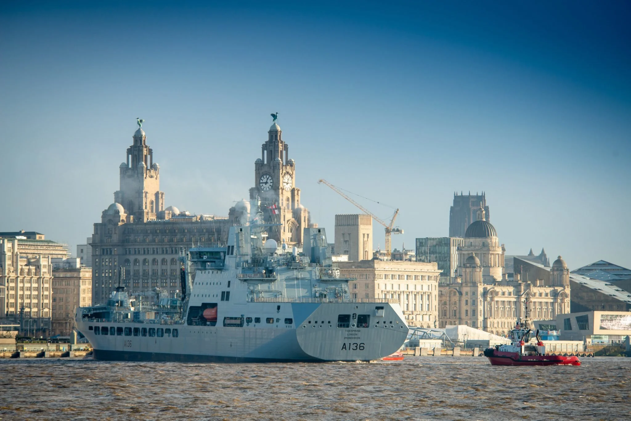 View of city skyline with historic buildings, a large ship in the water, and a smaller boat nearby under a clear blue sky.