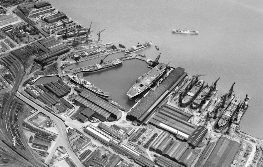 Aerial view of a shipyard with multiple ships docked, cranes, warehouses, and industrial buildings along the waterfront.