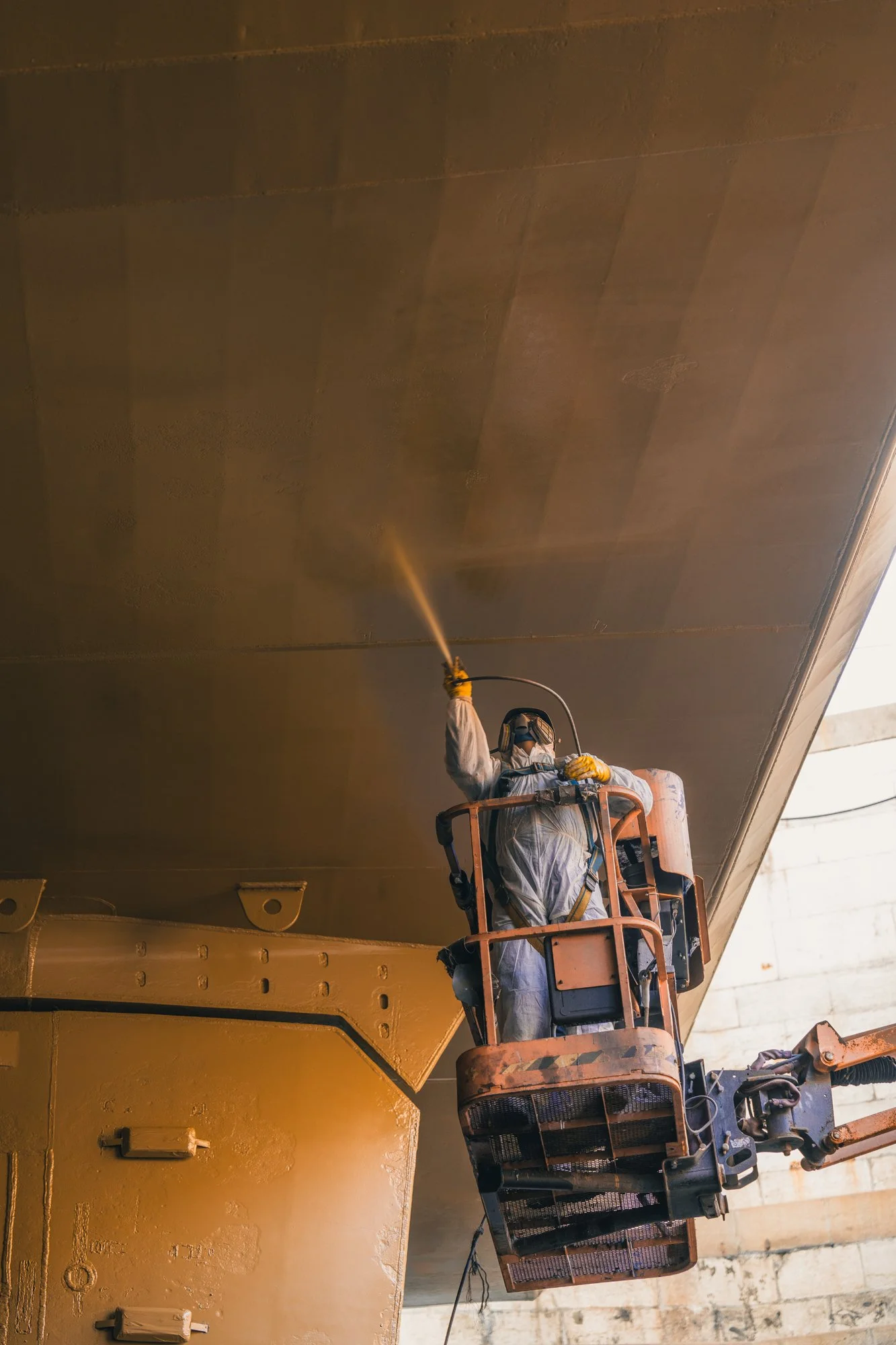 A worker wearing protective gear is on an elevated work platform applying a brown coating or paint to the underside of a large structure, possibly an aircraft or industrial equipment.