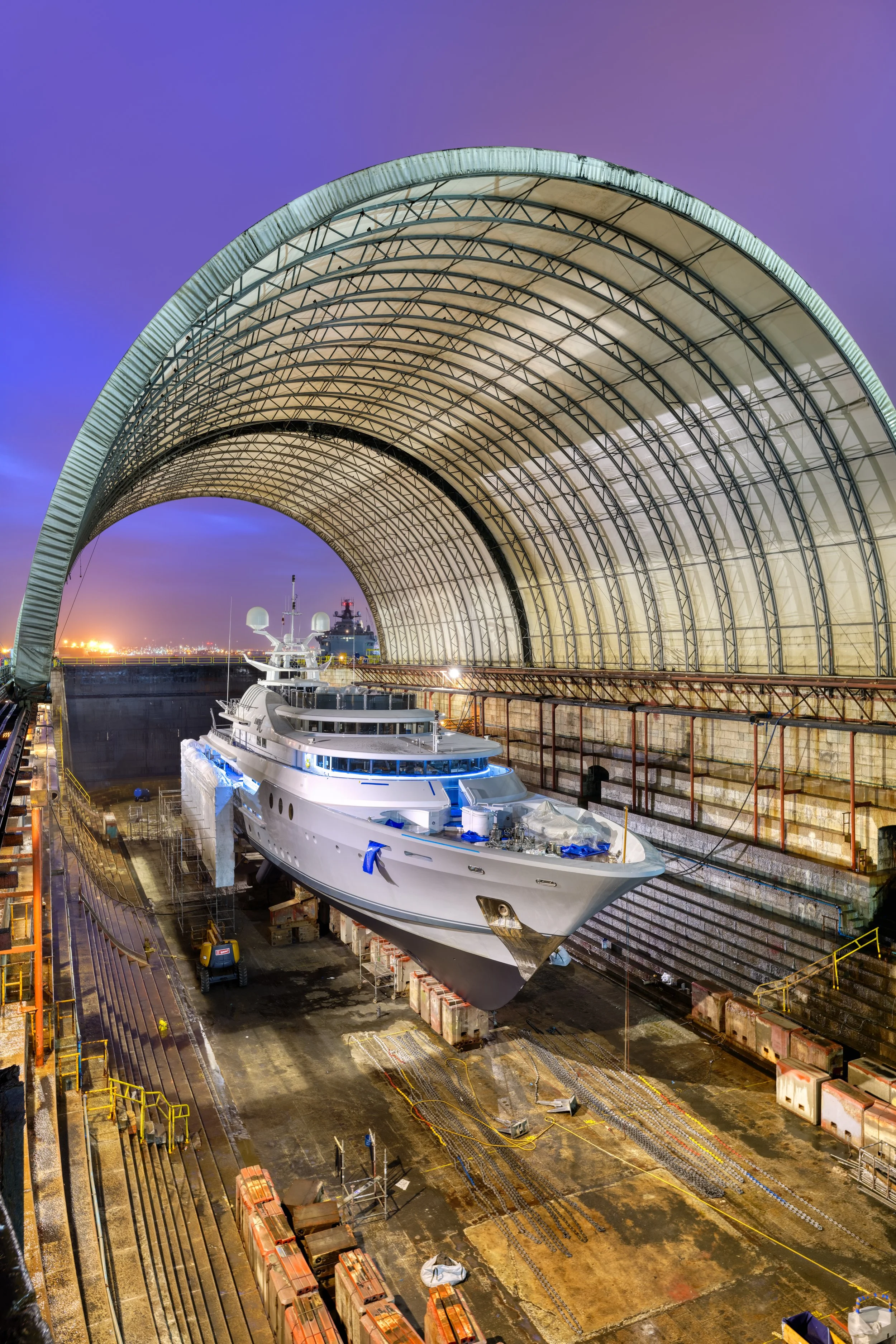 A large yacht inside a shipyard dry dock under a large curved metal roof at dusk, with city lights visible in the background.