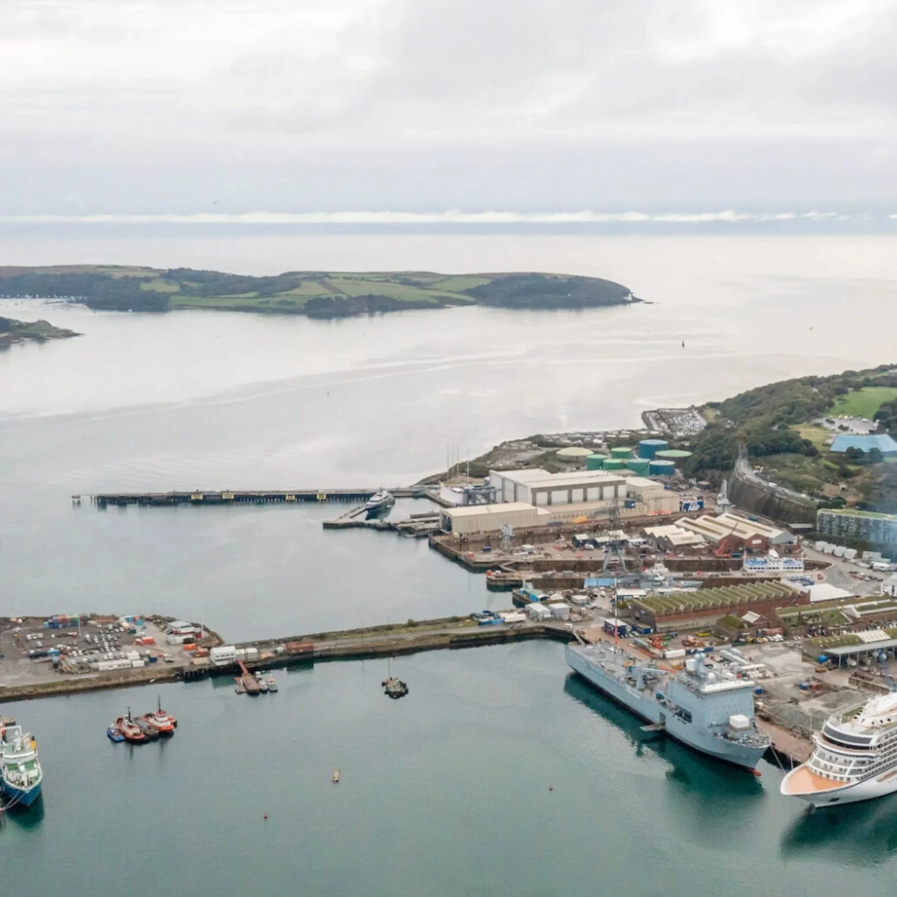 Aerial view of a harbor with ships and boats, industrial buildings, and a body of water with an island in the background.
