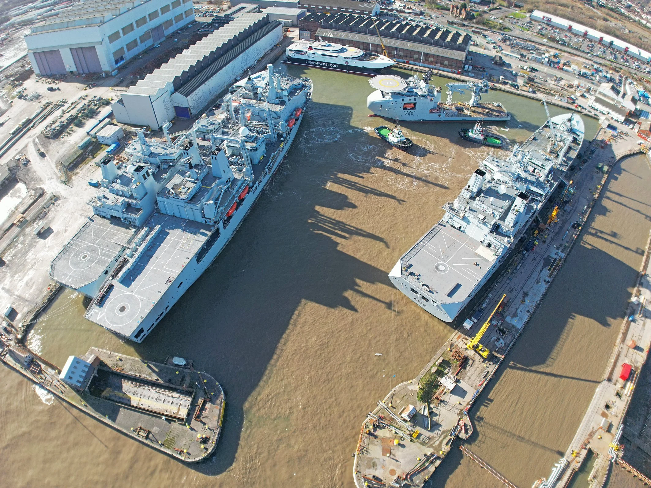 An aerial view of Balaena Birkenhead  with multiple ships docked next to large industrial buildings, a boat with a website URL, and surrounding parking lots and infrastructure.