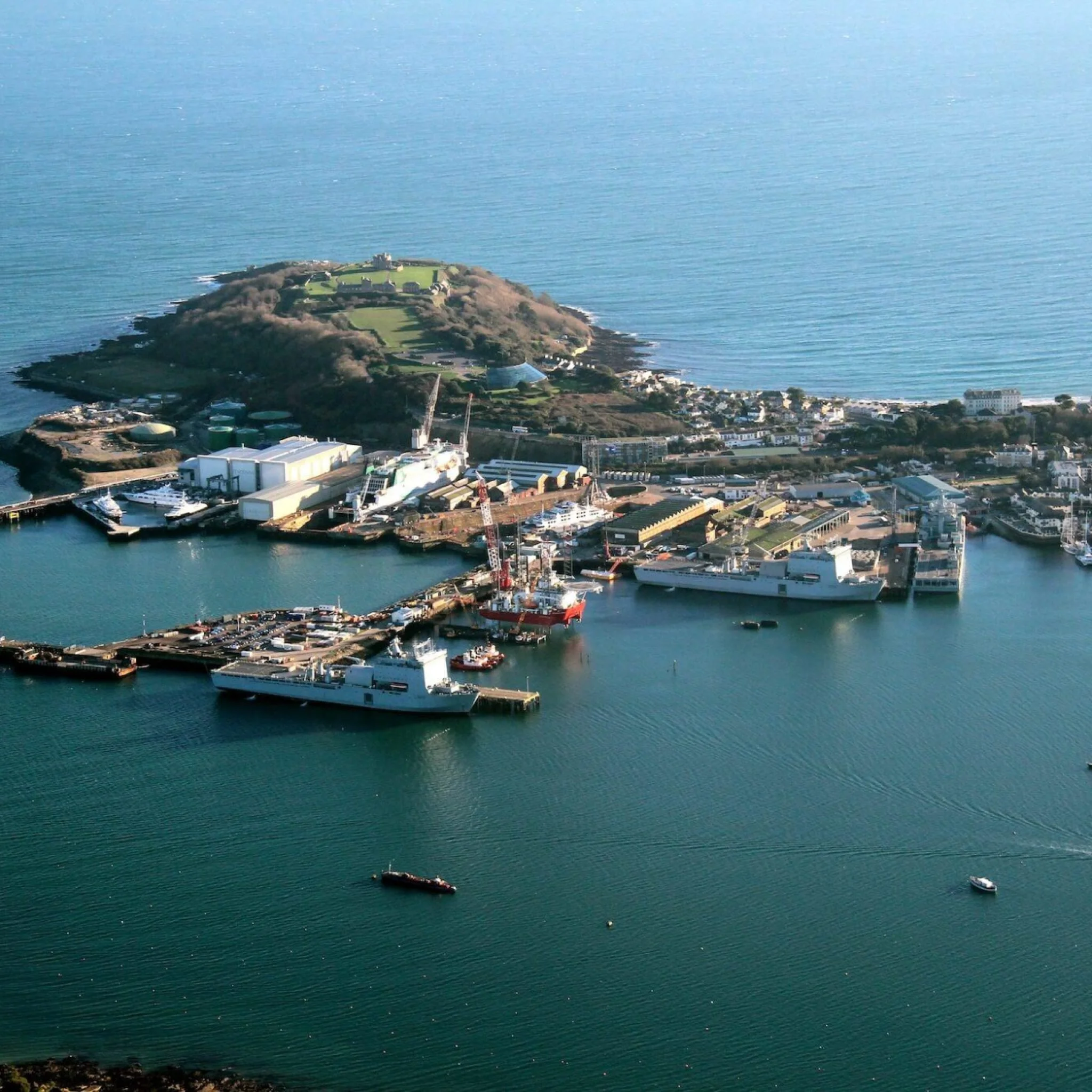 Aerial view of a harbor with ships docked, industrial buildings, and a lush green island in the background near the coastline.