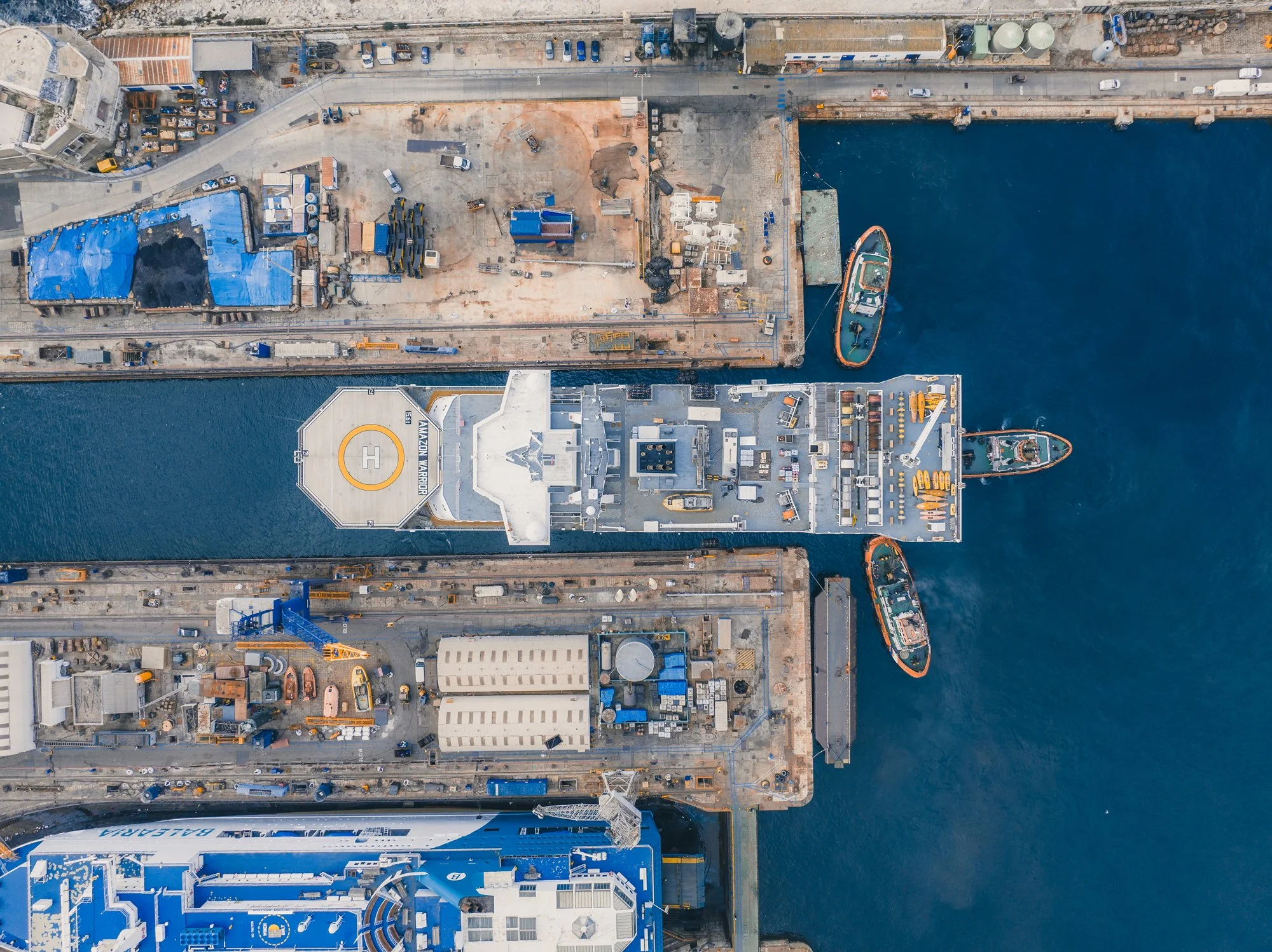 Aerial view of a construction site with equipment, vehicles, and materials near a body of water, adjacent to a large building with a helipad marked 'HELICOPTER'.