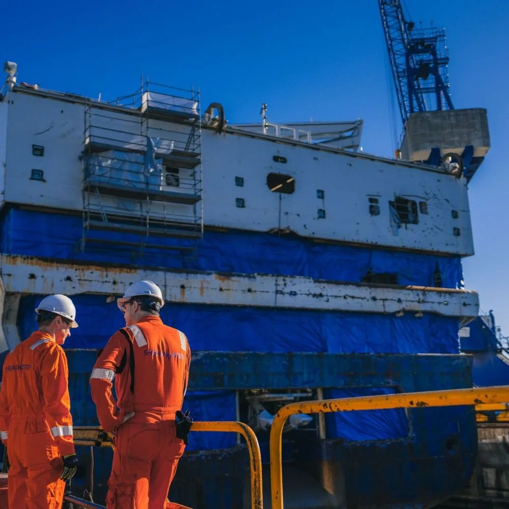 Two workers in orange safety gear and white helmets standing in front of a rusty, weathered ship under construction or repair at a dock with a large crane in the background.