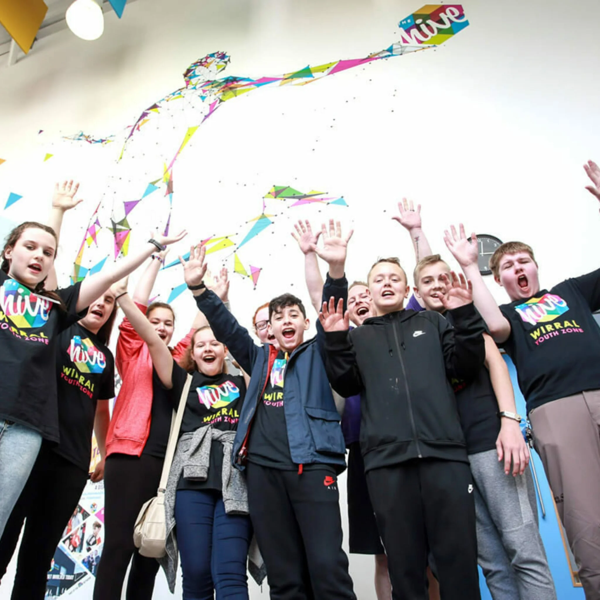 A group of people raising their hands and cheering at an indoor event with colorful banners and a mural of a running figure, indicating a lively and energetic atmosphere.