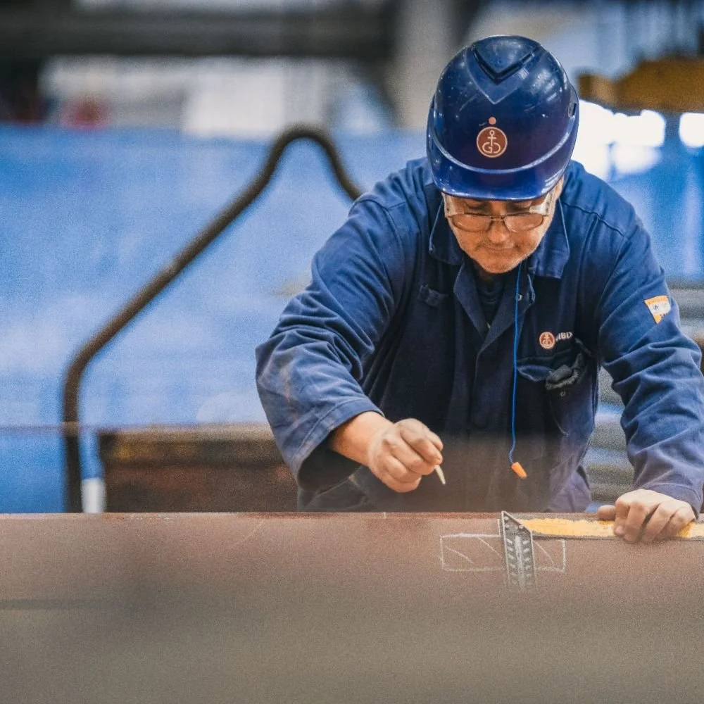 Worker in blue uniform and safety helmet drawing on a large metal surface in an industrial setting.