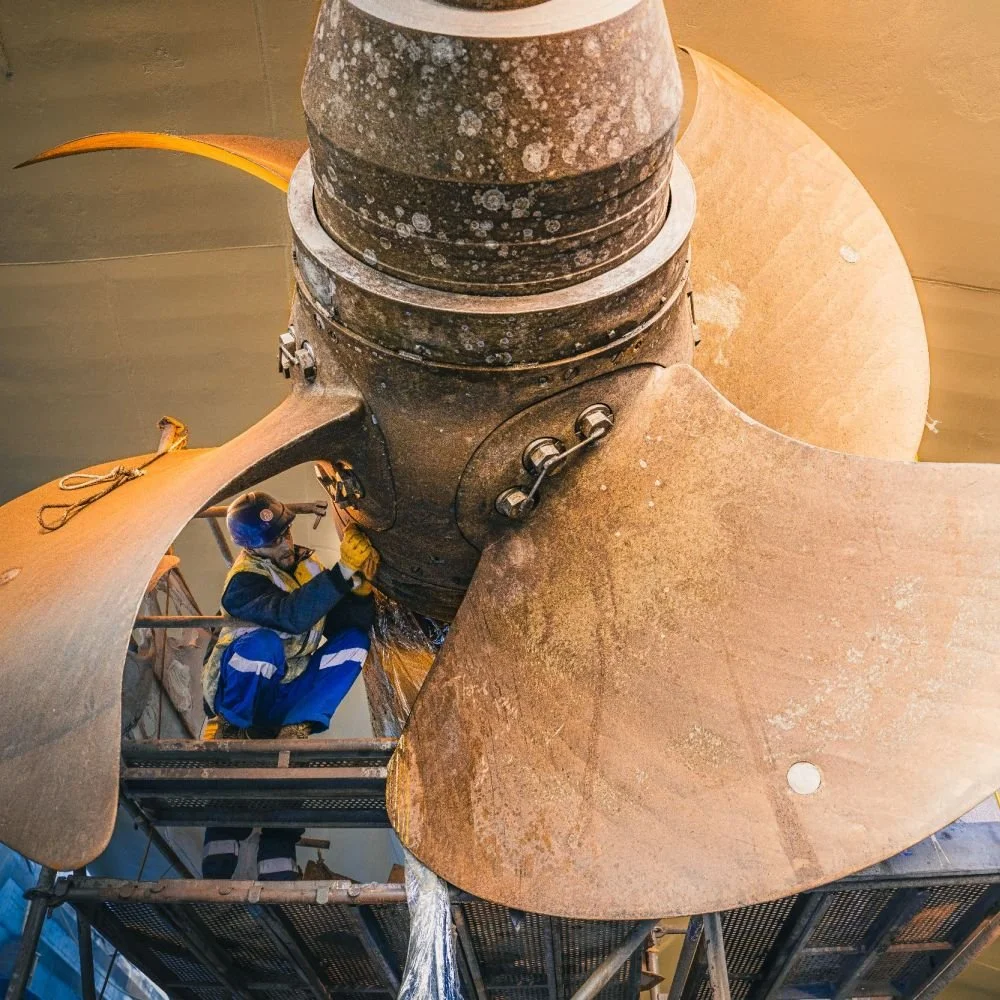 A worker in blue and yellow protective gear working on a large, industrial metal turbine or propeller blade, with scaffolding underneath.