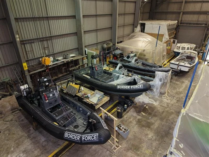 Several boats, including border patrol boats, are stored inside a large warehouse or hangar. Some boats are in various stages of assembly or repair.
