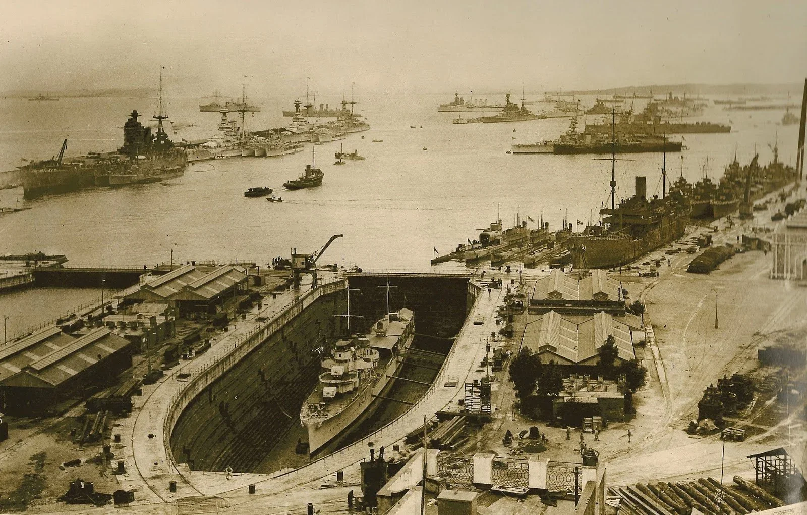 Black and white historical photograph of a busy naval shipyard with battleships docked at the quayside, several smaller boats in the water, and shipbuilding activity on land.