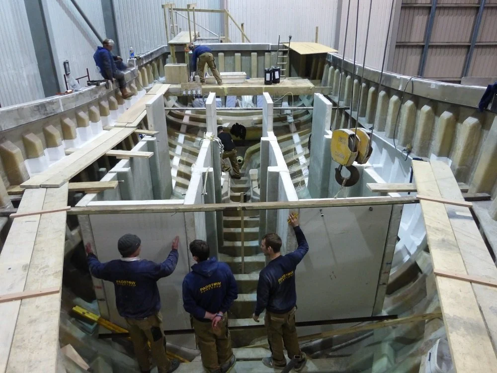 Construction workers building a large indoor structure with concrete and steel, possibly a swimming pool or water feature, inside a warehouse.