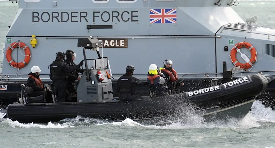 Border Force boat with four officers, some wearing helmets and life jackets, on the water near a larger vessel marked 'Border Force' and a UK flag.