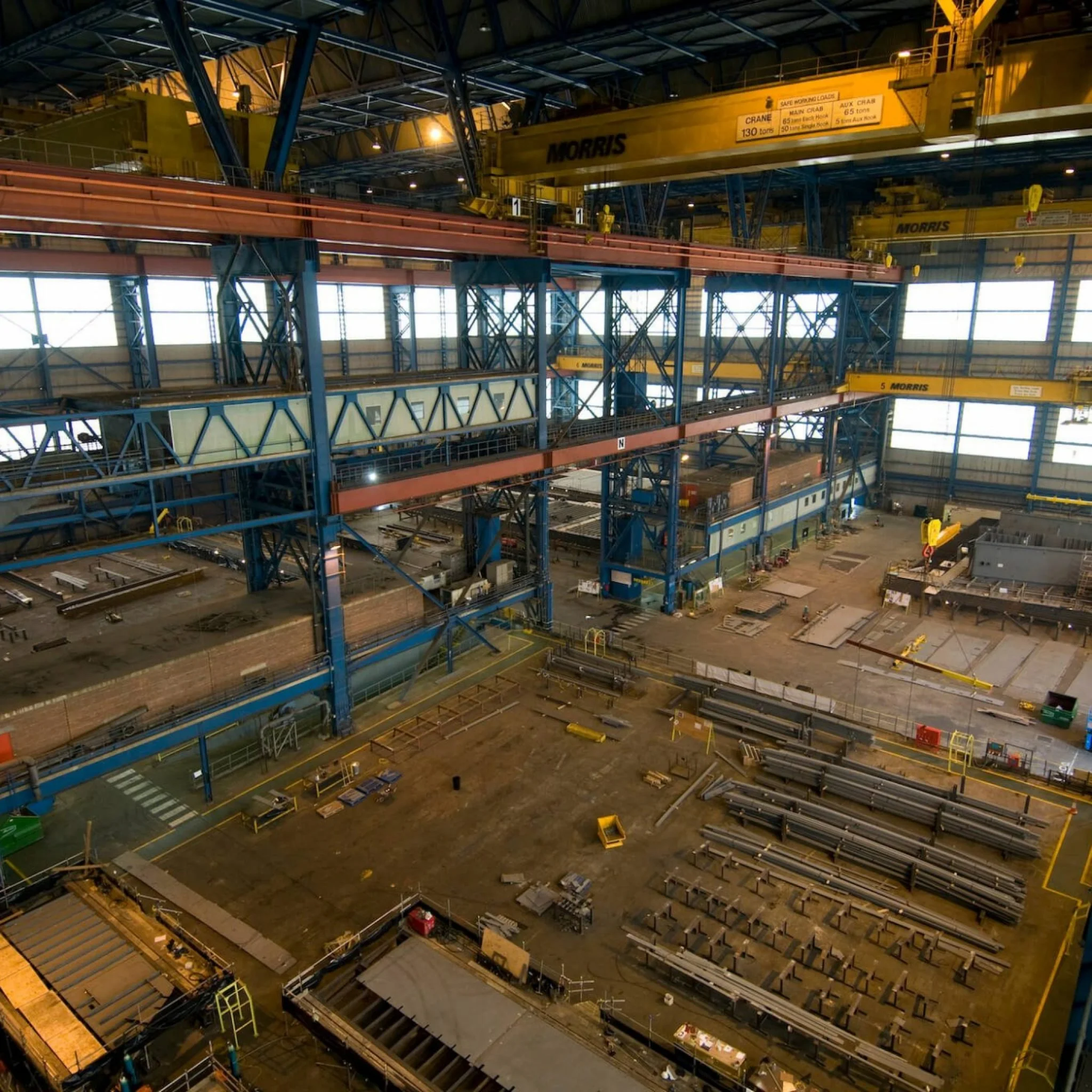 Inside view of a large industrial warehouse with high ceilings, blue and yellow overhead cranes, and various metal materials and equipment scattered on the floor.