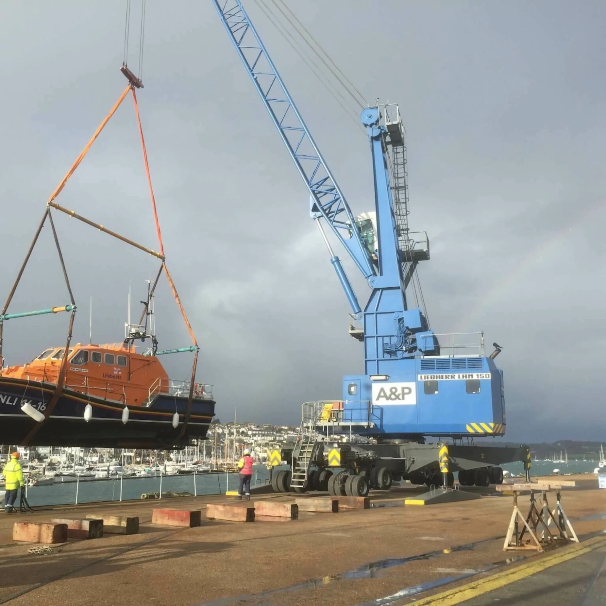 A large blue crane lifting a bright orange lifeboat at a harbor with sailboats and buildings in the background.