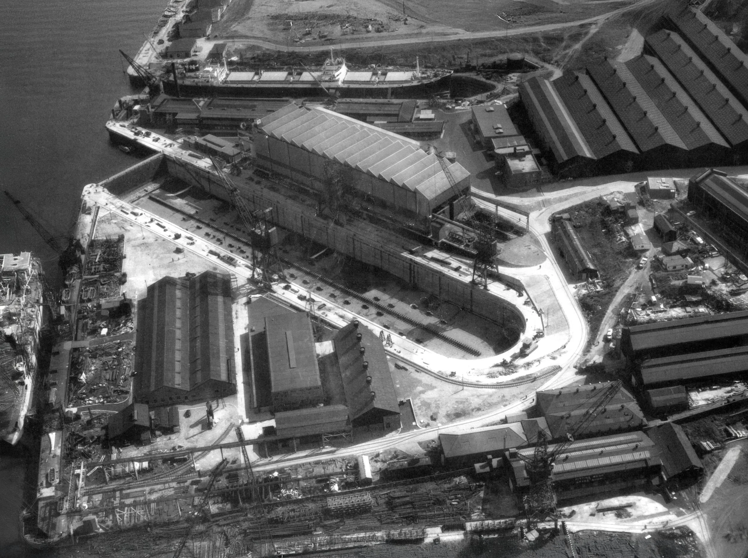 Black and white aerial photograph of a construction site with cranes, buildings, and a large ship docked near the water's edge.