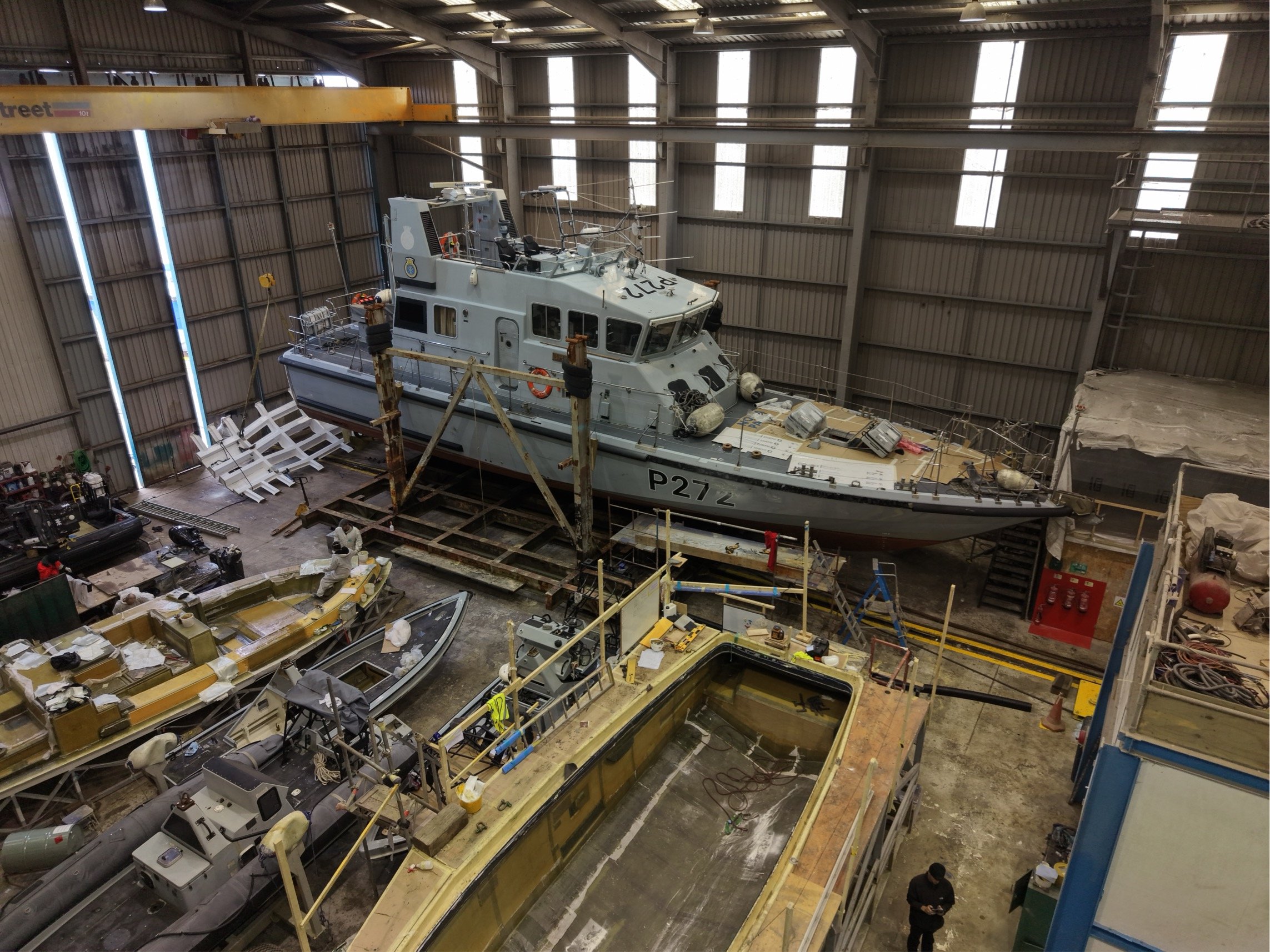 Photo of an indoor boat manufacturing or repair facility featuring a large gray boat with the registration P27Z inside a spacious warehouse with high ceilings and multiple windows, surrounded by machinery, tools, and smaller boats in the workshop.