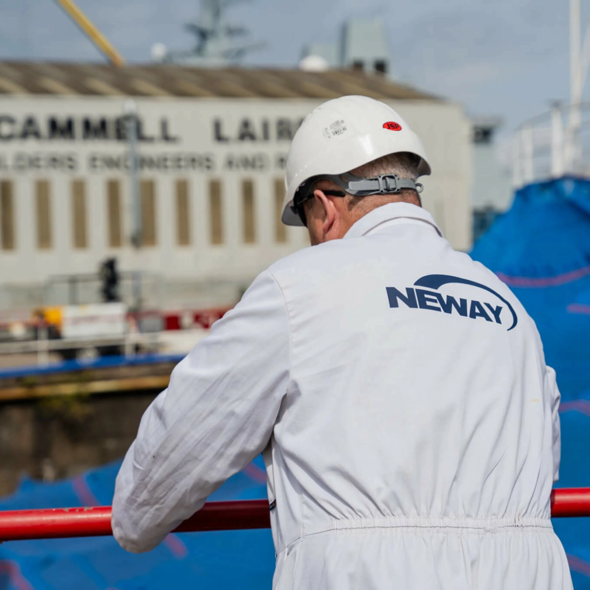 A person wearing a white safety helmet and coveralls with the word 'NEWAY' on the back, leaning on a red railing near a dock or industrial site.