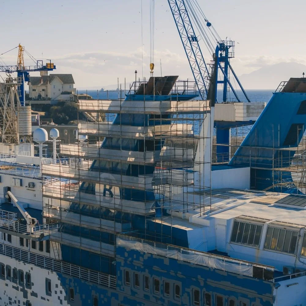 A cruise ship docked at a harbor under scaffolding with construction equipment, cranes, and buildings visible in the background.