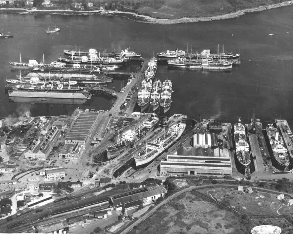 Aerial black and white photo of a harbor with several ships docked, buildings, and surrounding land.