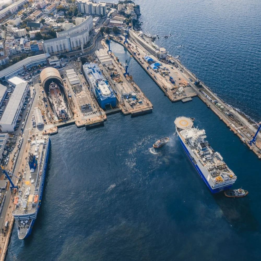 Aerial view of a shipyard with construction and maintenance facilities, two large ships docked, a small boat moving nearby, and a coastal city in the background.
