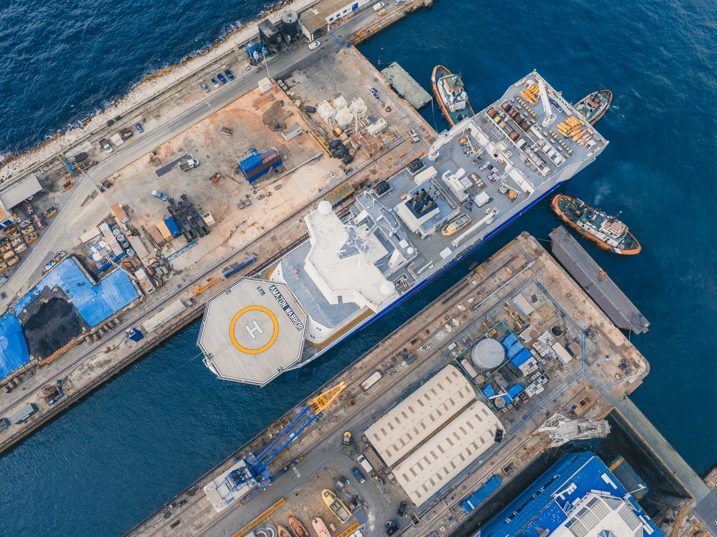 Aerial view of a harbor with ships docked, including a large vessel with a helipad and helipad markings, surrounded by industrial buildings, equipment, and parked vehicles.