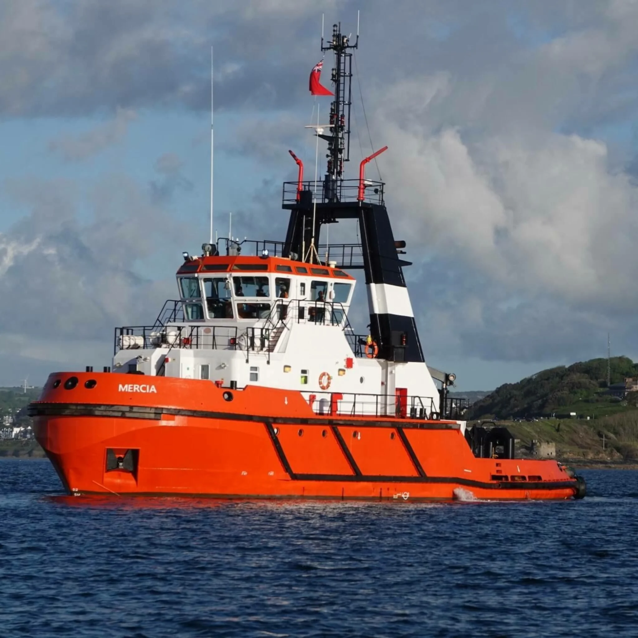 A bright orange and white tugboat named Mercia sailing on water with a hilly shoreline in the background and cloudy sky overhead.