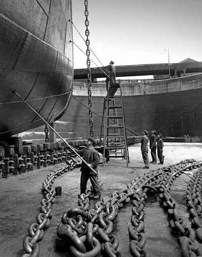Shipyard workers installing large chains on a ship using a ladder and crane in black and white.