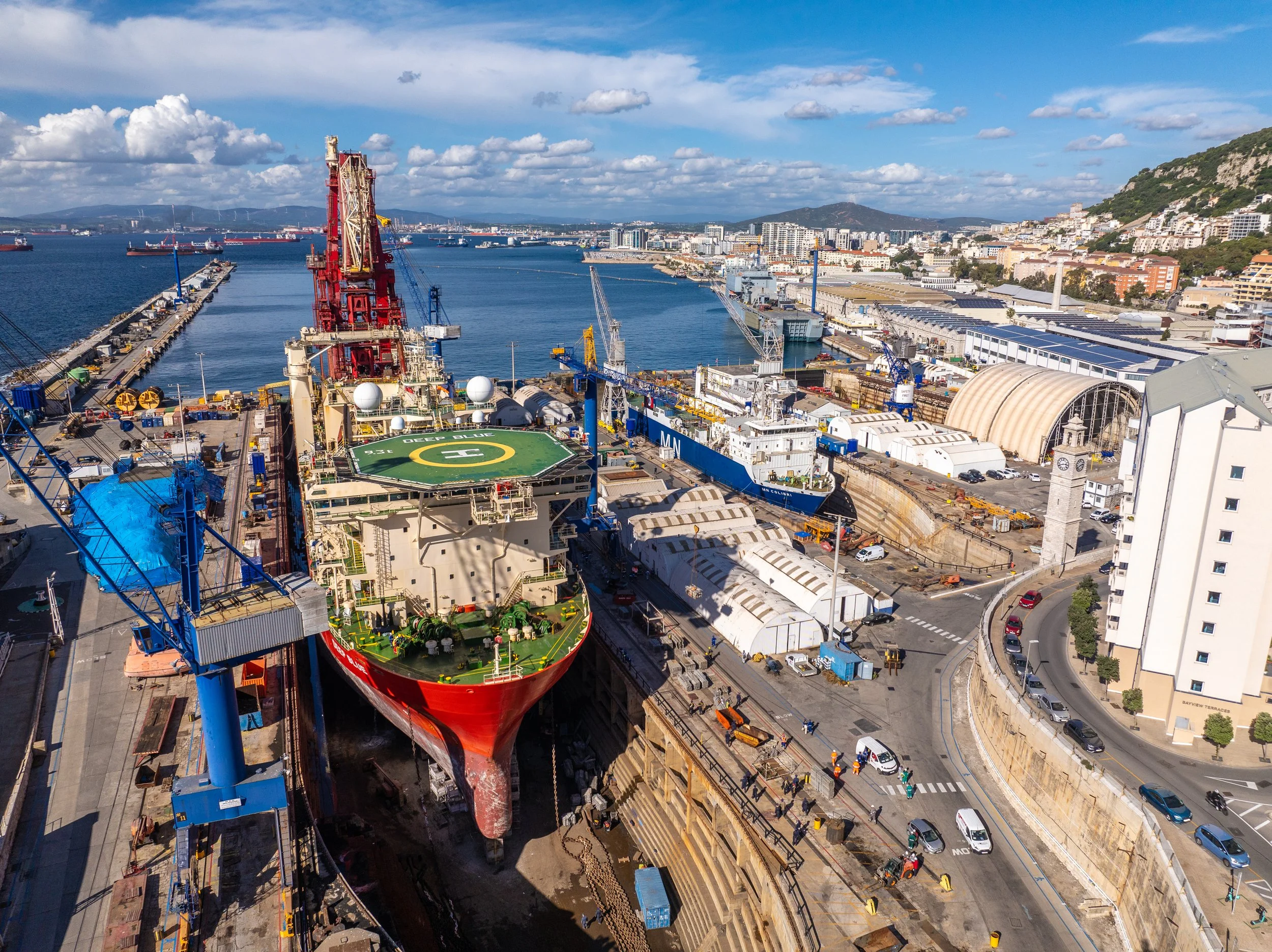 A large dock with a red and green ship undergoing construction or maintenance, surrounded by cranes and equipment, with a cityscape and water in the background, under a partly cloudy sky.