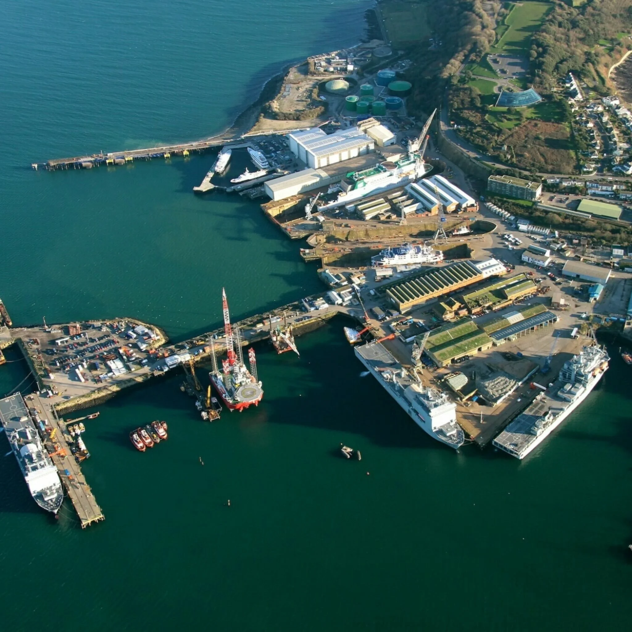 Aerial view of a harbor with boats, industrial buildings, and a marina on the water, with roads and greenery surrounding the area.