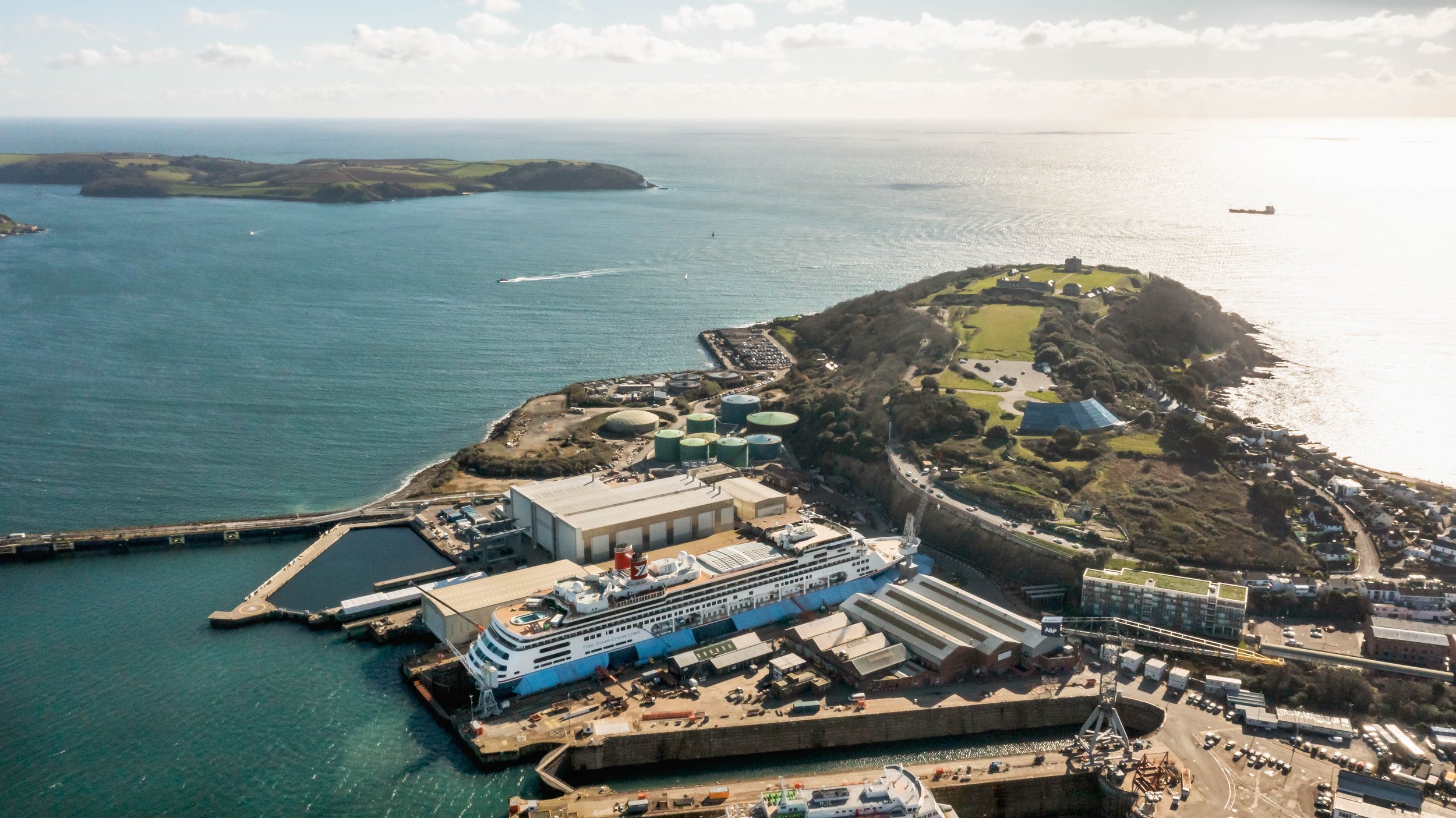 Aerial view of Balaena Falmouth with a cruise ship docked, surrounded by industrial buildings, green hills, and water with boats, under a partly cloudy sky.