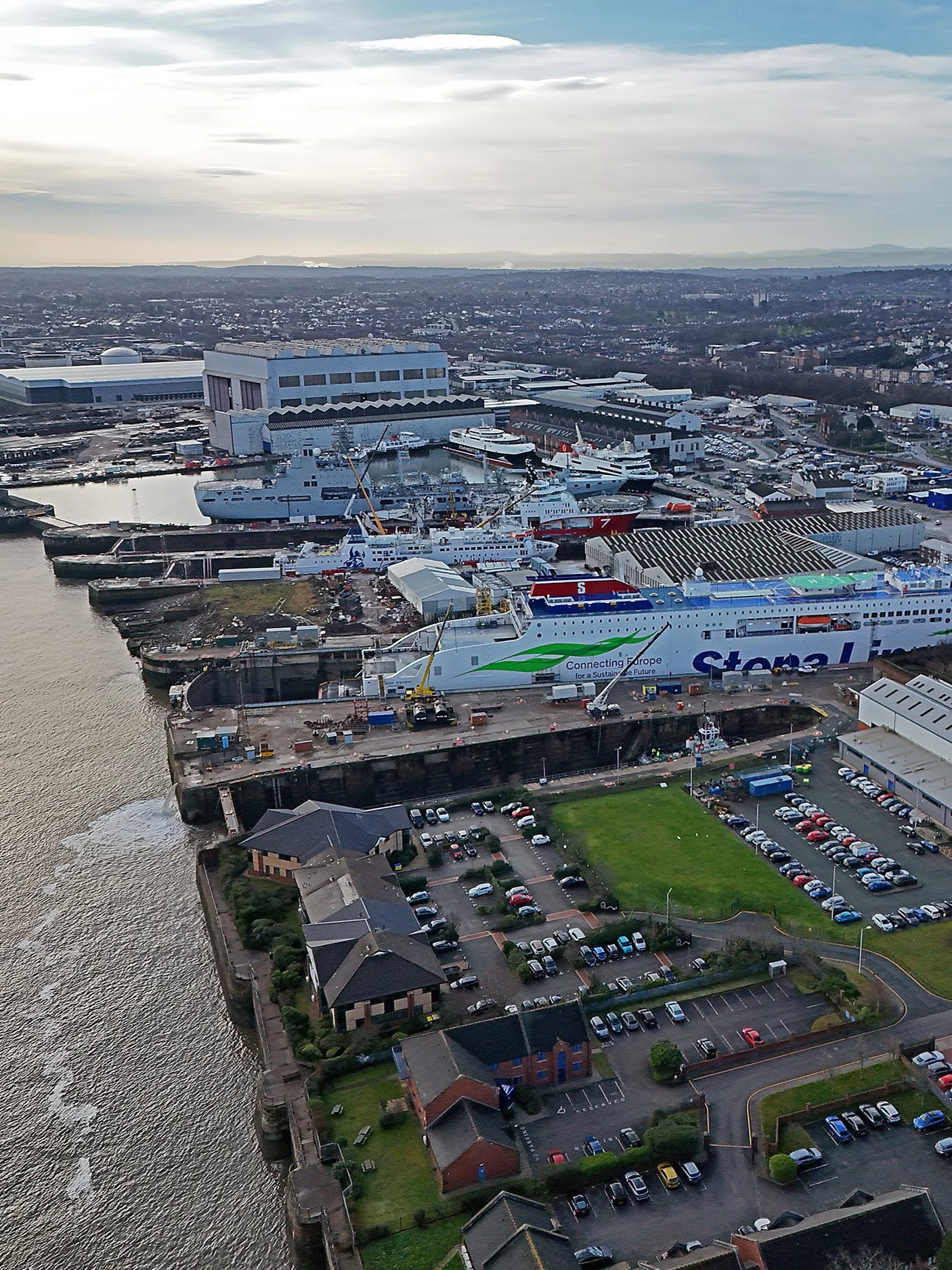 Aerial view of Balaena Birkenhead with ships, industrial buildings, parking lots, and residential houses near water under a cloudy sky.